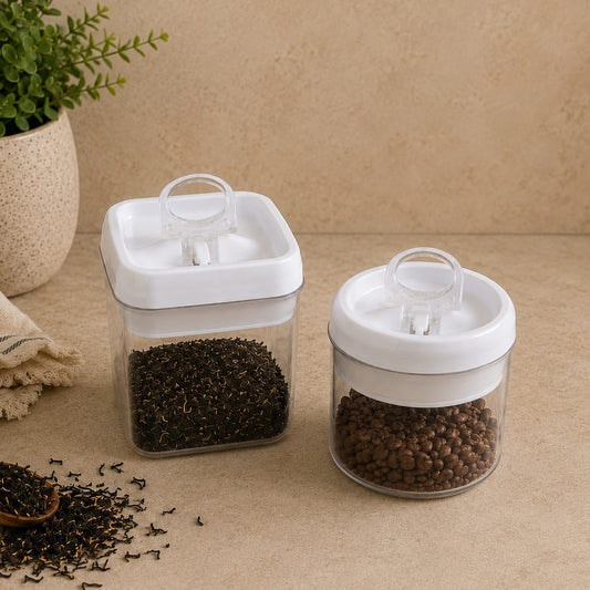 Two clear airtight plastic spice jars with white lids and clear flip-top dispensers, filled with dark tea and brown peppercorns, set on a beige countertop with a potted plant and cloth in the background.